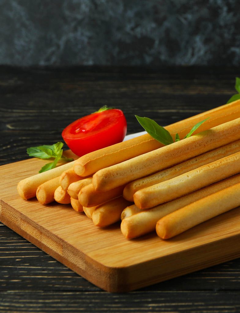 Grissini dorati e croccanti su tagliere di legno con pomodoro fresco, snack da forno ideale per aperitivi e antipasti.