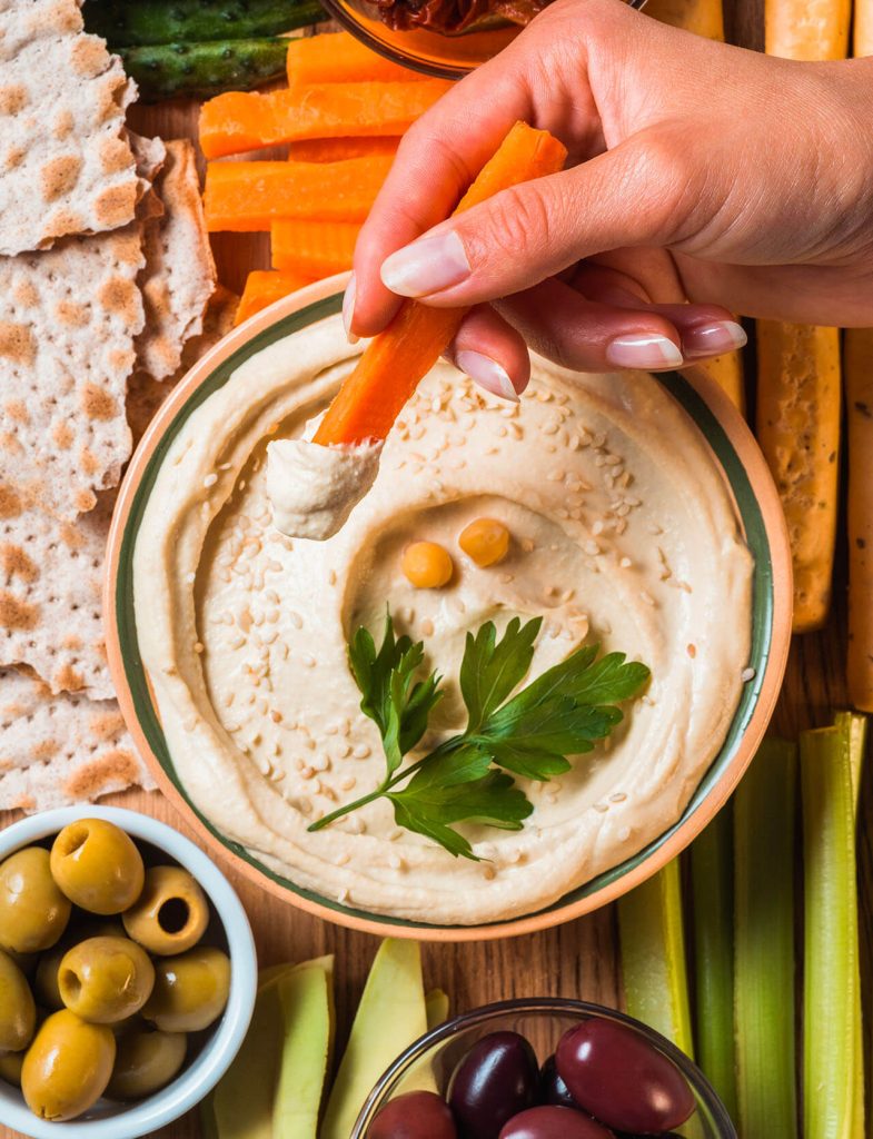 Hummus cremoso decorato con ceci e prezzemolo, servito con verdure fresche, olive e pane croccante.