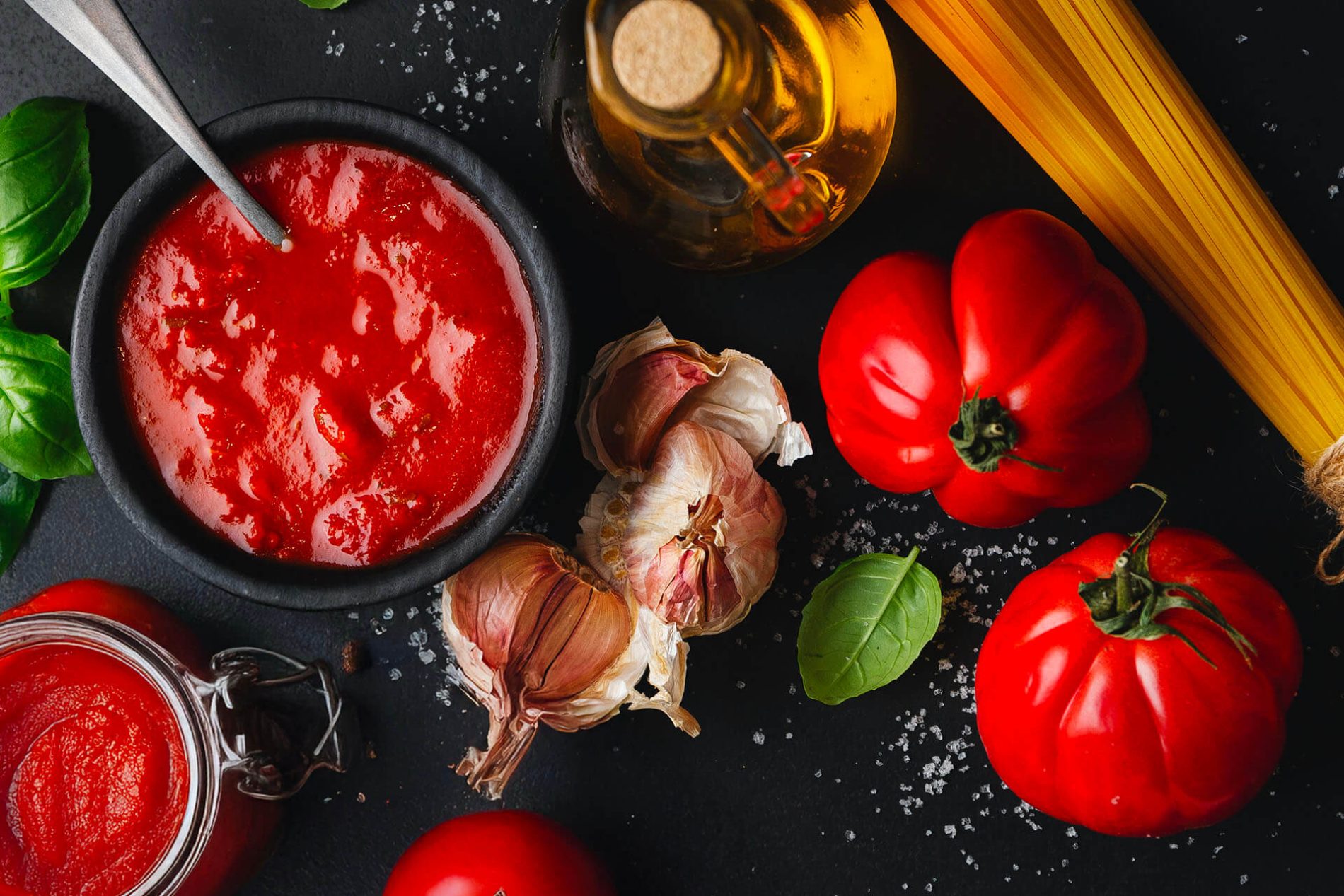 Pomodori e Sughi — macro: ciotola di passata con pomodori freschi, aglio, basilico e spaghetti sullo sfondo