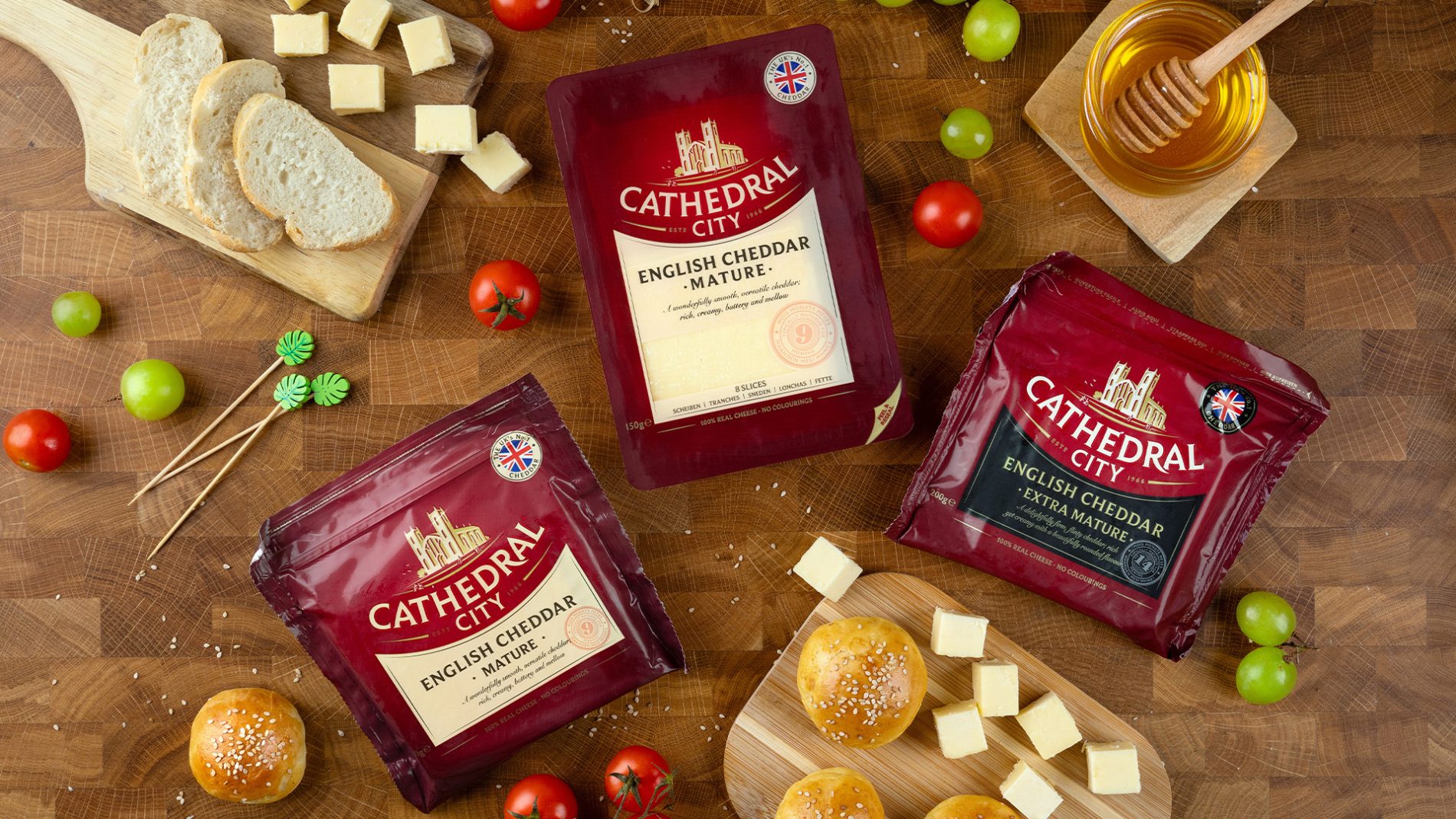Confezioni di cheddar inglese Cathedral City Mature ed Extra Mature, distribuito in Italia da Atlante, con formaggio e ingredienti su un piano in legno.