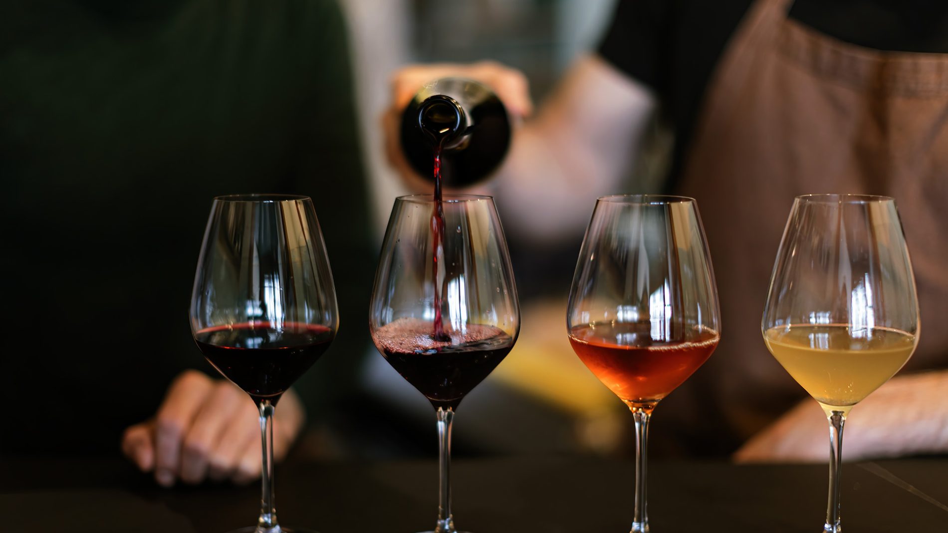 Primo piano di quattro calici di vino disposti in fila: due rossi, un rosato e un bianco. Una persona, sfocata sullo sfondo, sta versando del vino rosso nel secondo bicchiere. Atmosfera da degustazione professionale, con focus sui colori e sulle texture dei vini nei calici.