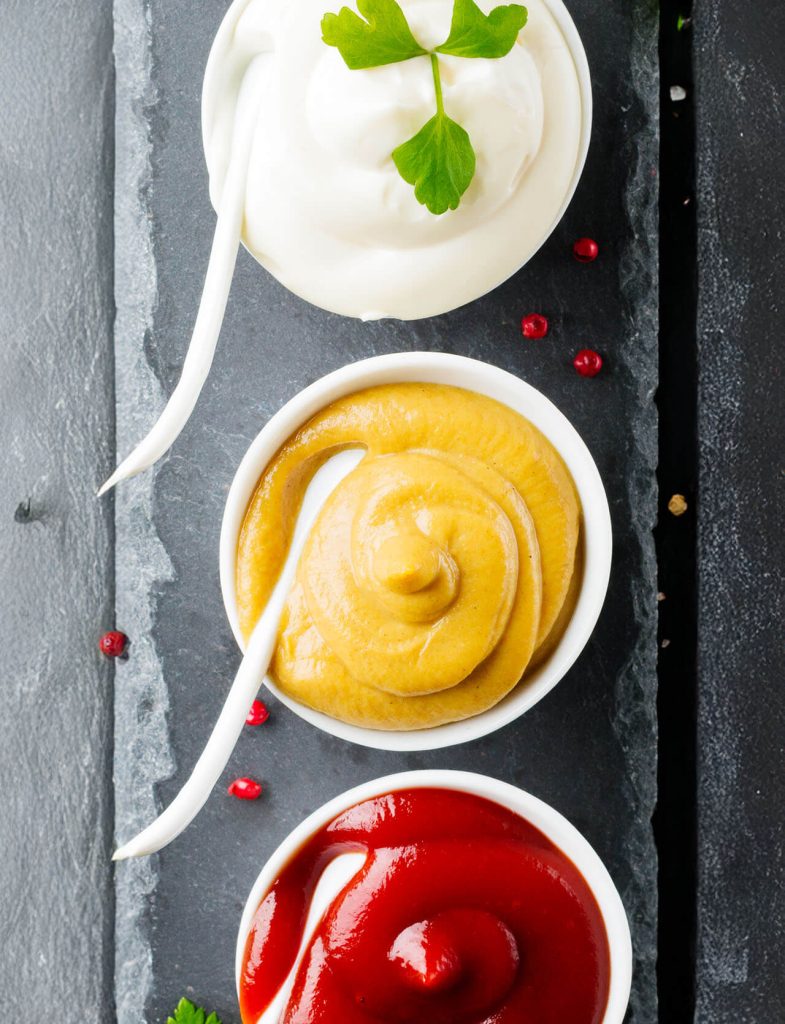 Maionese, ketchup e senape in ciotole di ceramica, disposte con erbe e spezie per condimenti classici.